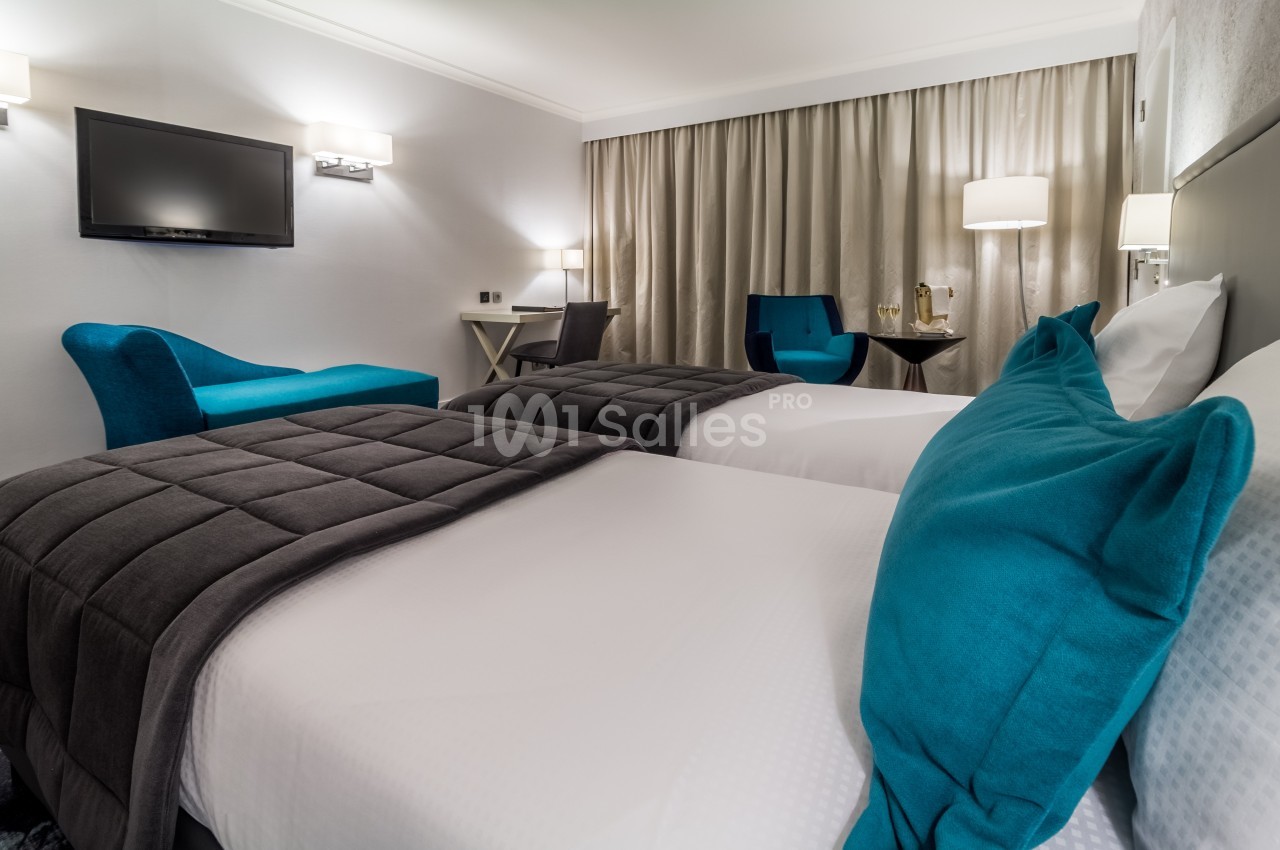Chambre d'hôtel avec deux lits simples, fauteuils bleus, bureau, rideaux beiges et éclairage doux.