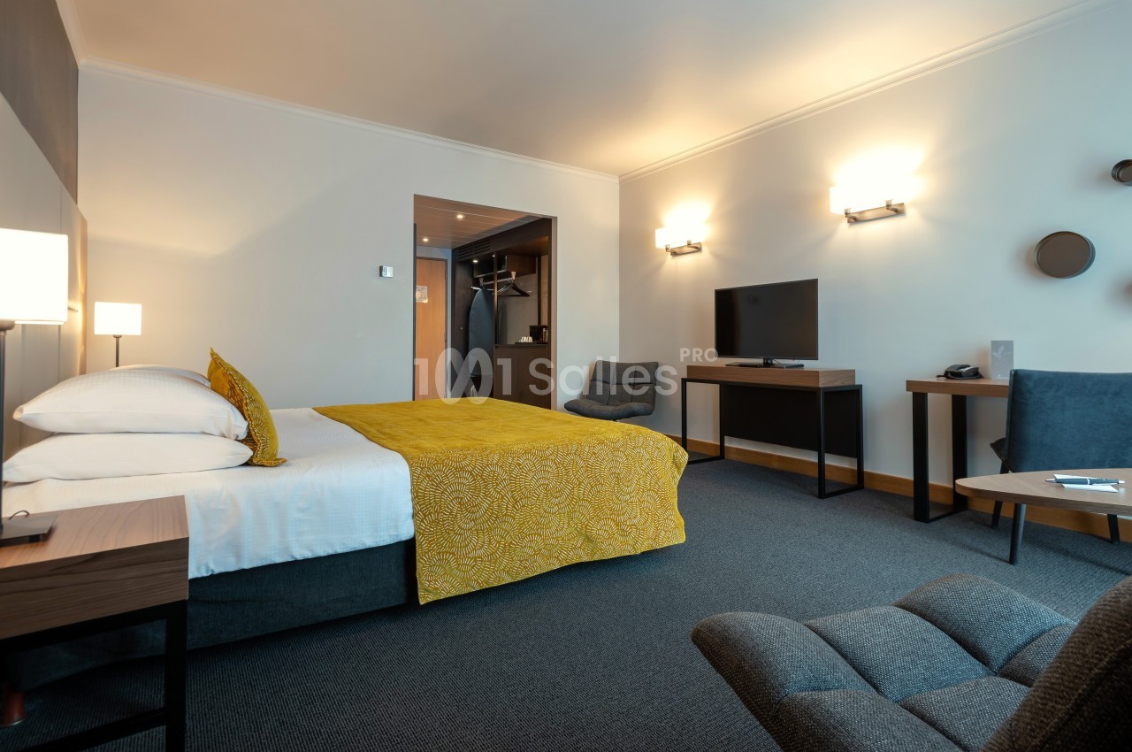 Chambre d'hôtel moderne avec lit double, bureau, télévision, fauteuils et éclairage chaleureux.