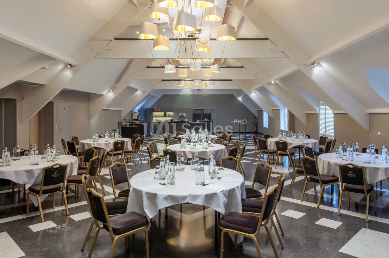 Salle de réception lumineuse avec tables rondes dressées, chaises disposées autour et un buffet en arrière-plan.
