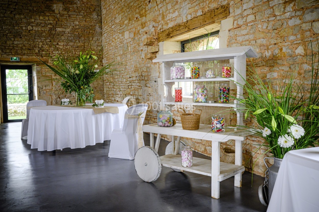Salle en pierre aménagée pour un événement, avec tables blanches et un chariot décoré de bonbons colorés.