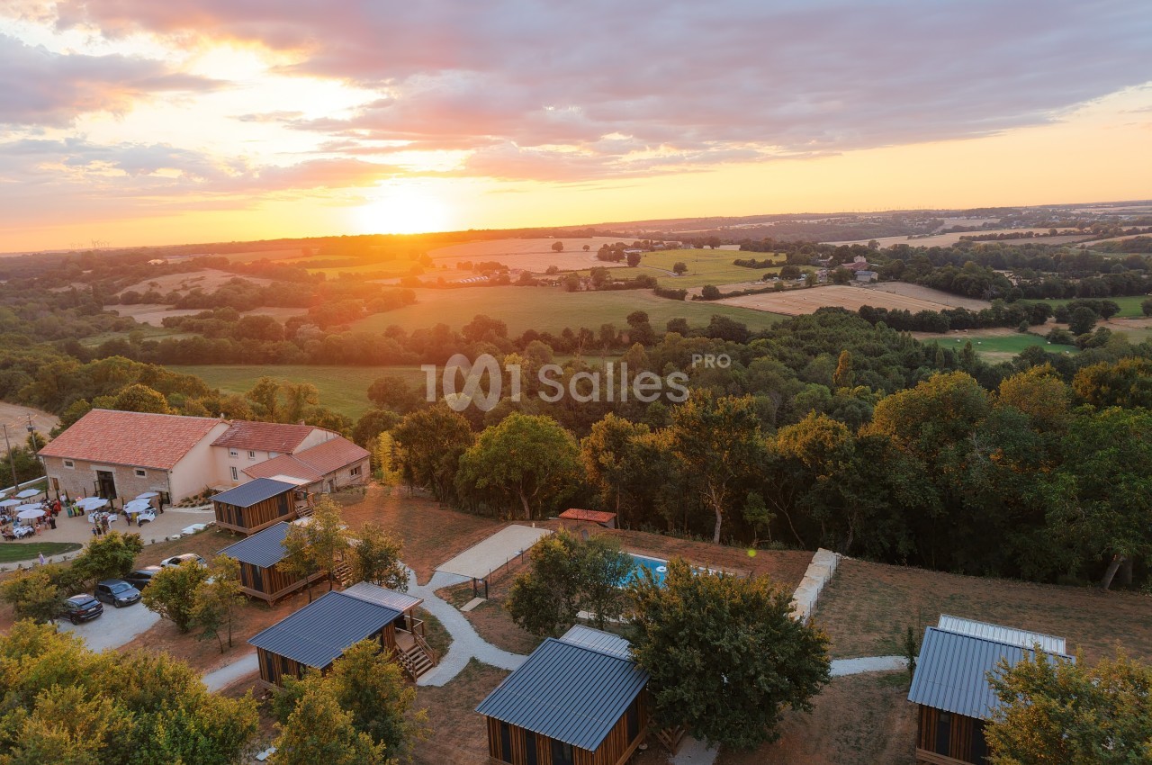 Vue aérienne d'un domaine rural au coucher du soleil avec chalets en bois, piscine et paysage vallonné.