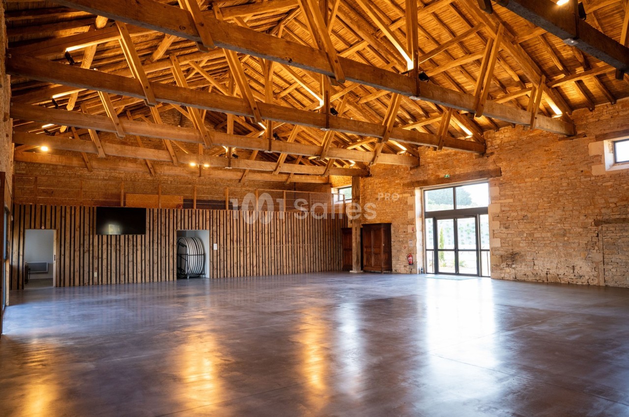 Intérieur d'une grande salle avec charpente en bois apparente, murs en pierre et sol en béton poli.