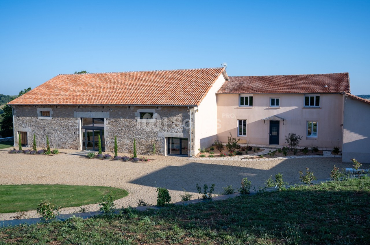 Bâtiment rénové en pierre et crépi avec toit en tuiles, entouré d'une cour gravillonnée et d'espaces verts.