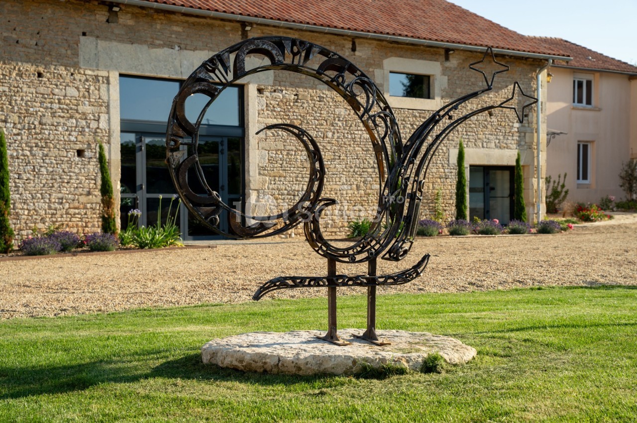Sculpture métallique en forme de coq stylisé sur une pelouse devant un bâtiment en pierre avec des fenêtres modernes.