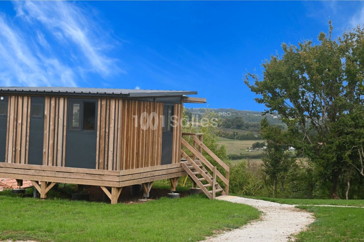 Cabane en bois surélevée avec terrasse, située dans un paysage rural verdoyant sous un ciel bleu.