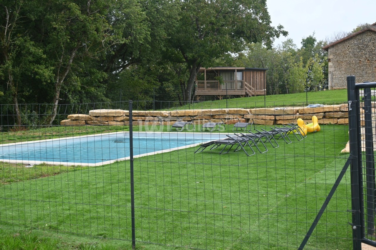 Piscine extérieure entourée de chaises longues sur pelouse, avec clôture et cabane en bois en arrière-plan.