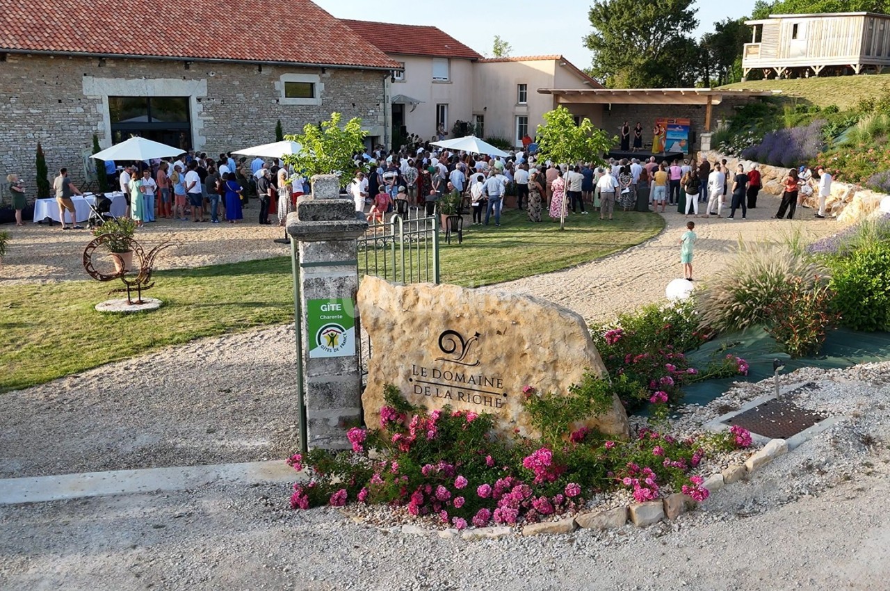 Vue d'un domaine avec des invités rassemblés en extérieur près d'un bâtiment en pierre et d'une enseigne fleurie.