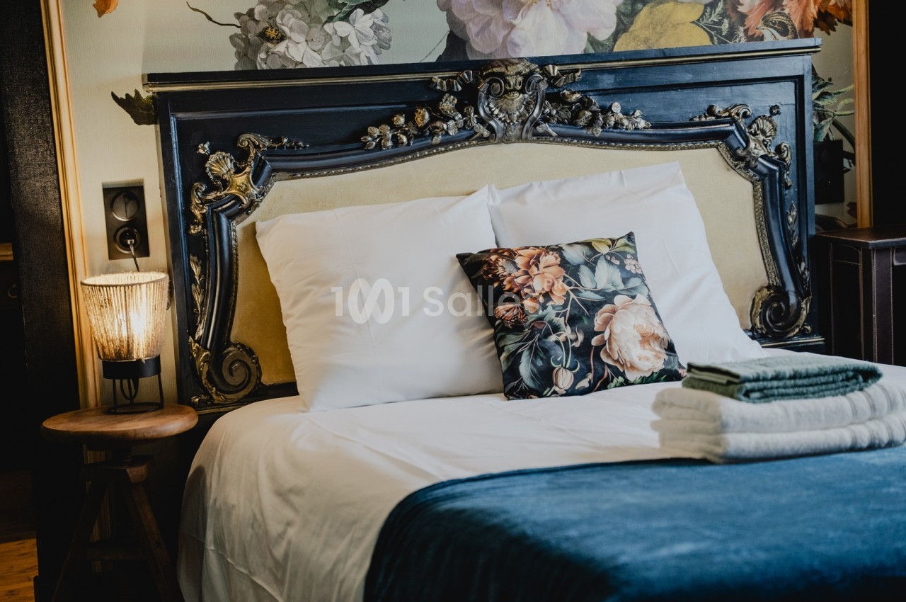 Lit avec tête de lit ornée, coussin à motif floral, lampe allumée sur une table en bois et serviettes pliées.