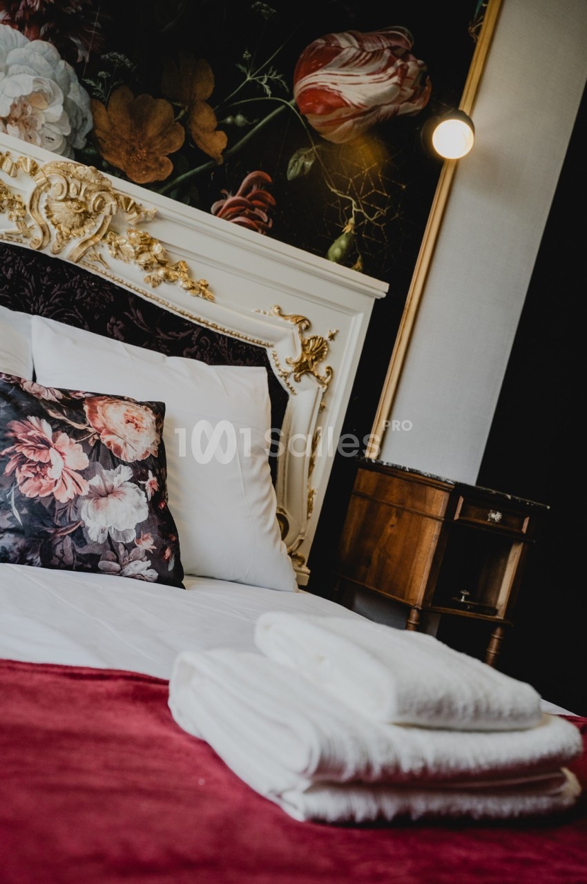 Lit avec tête de lit ornée de dorures, coussins fleuris, draps blancs et serviettes pliées sur une couverture rouge.