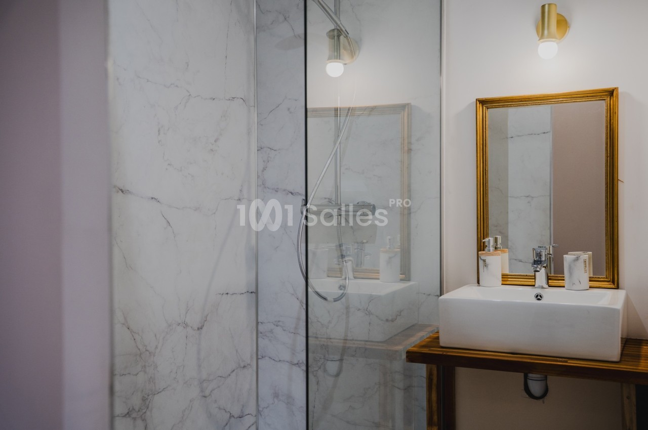 Salle de bain avec douche en verre, lavabo blanc sur meuble en bois et miroir encadré doré.