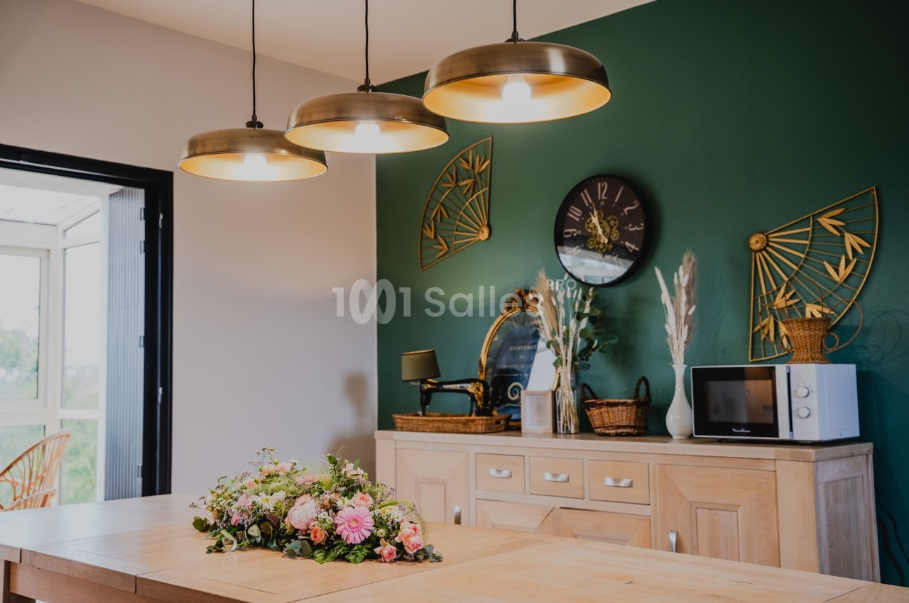 Suspension lumineuse au-dessus d'un îlot central avec bouquet floral, mur vert décoré et mobilier en bois clair.