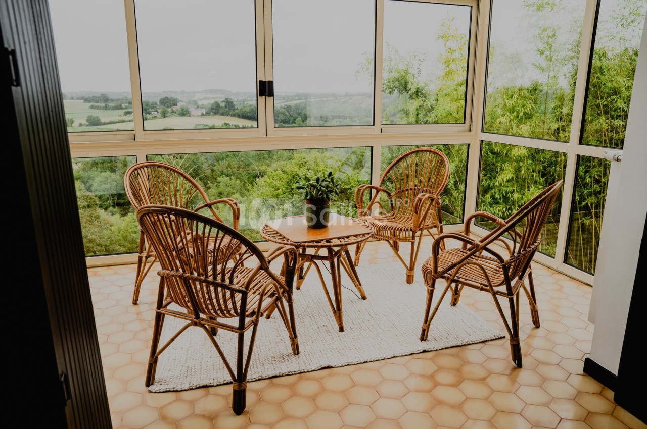 Quatre fauteuils en rotin autour d'une table basse sur un tapis clair dans une véranda avec vue sur la campagne.