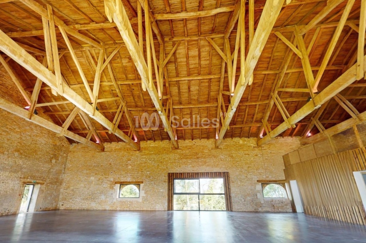 Charpente en bois apparente dans une grande salle aux murs en pierre, avec des fenêtres laissant entrer la lumière naturelle.