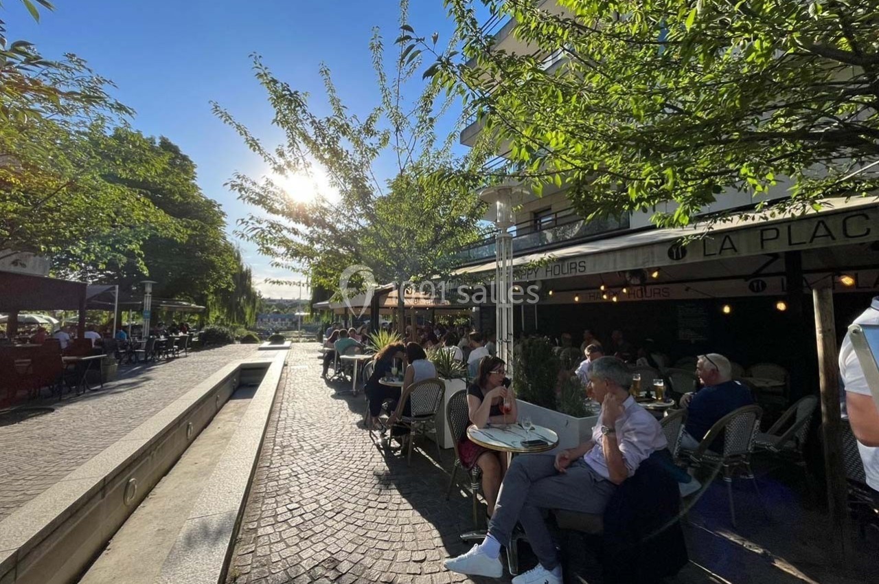 Terrasse de café animée sous des arbres, avec des clients attablés et une lumière de fin d'après-midi ensoleillée.