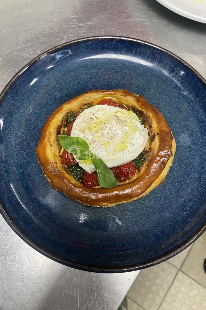Tartelette garnie de tomates confites, pesto, œuf poché et feuille de basilic, servie dans une assiette bleue.