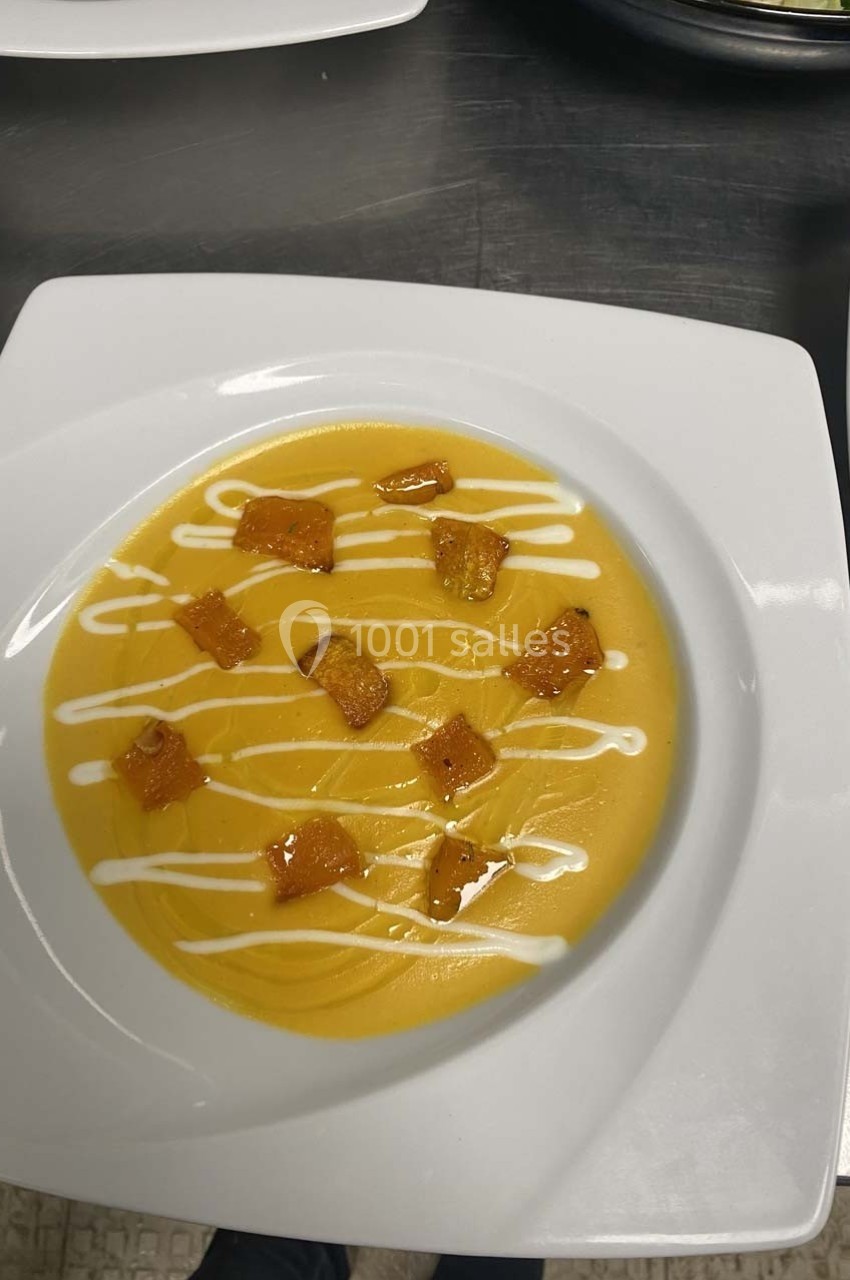 Assiette de velouté orange garnie de croûtons dorés et de traits de crème blanche, servie sur une table en inox.