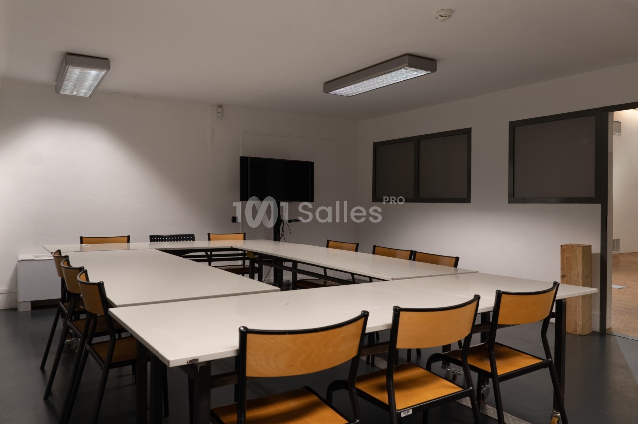 Salle de réunion vide avec une grande table en U, des chaises en bois et un écran noir fixé au mur.