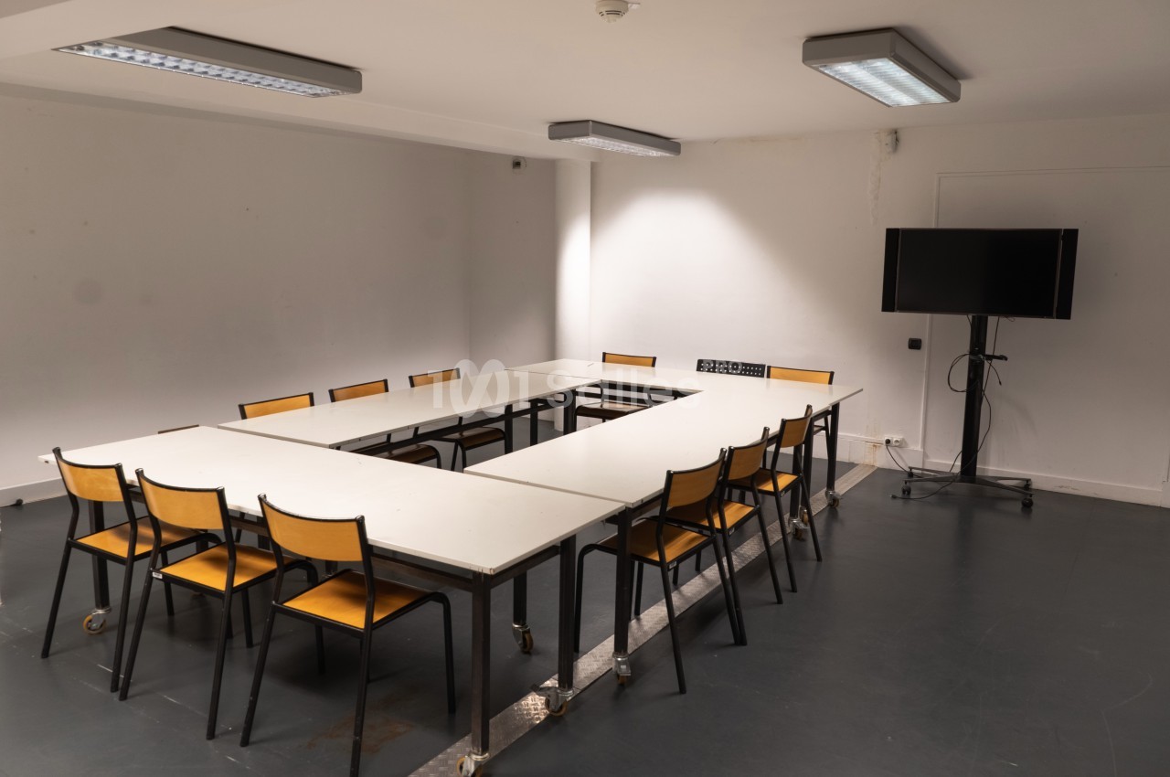Salle de réunion vide avec des tables disposées en U, des chaises en bois et un écran noir sur pied.