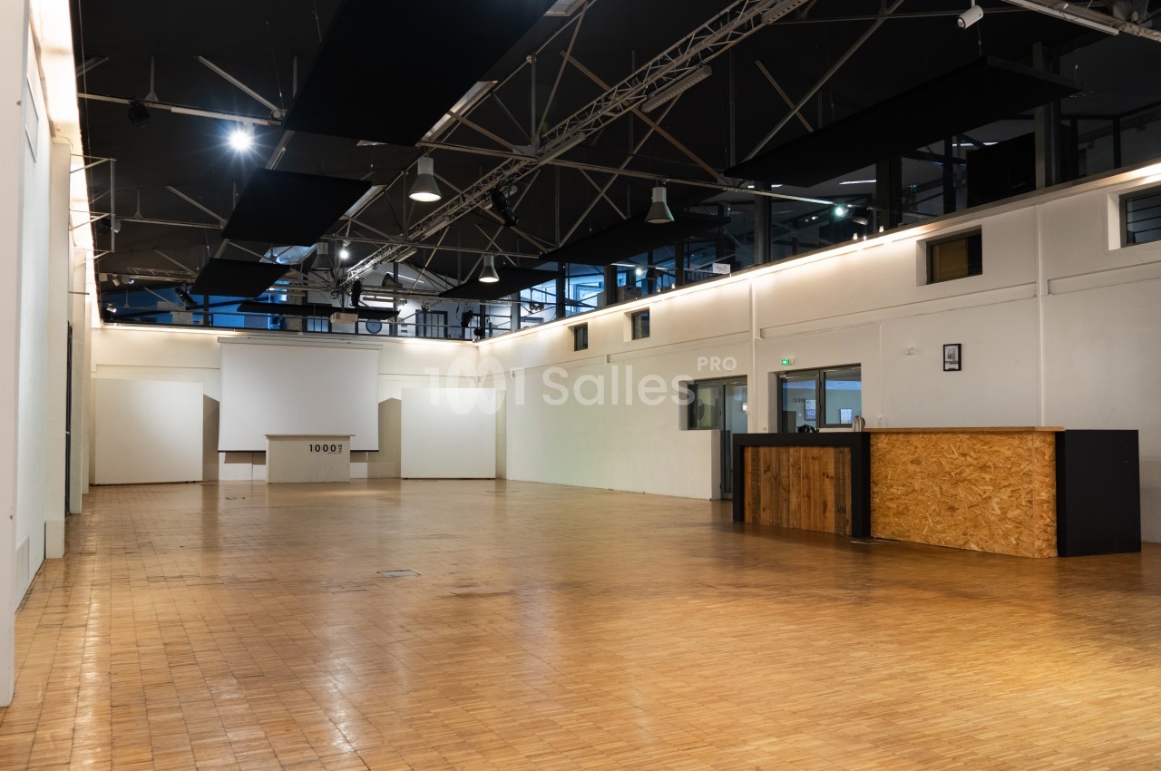 Salle spacieuse avec parquet en bois, murs blancs, plafond industriel et comptoir en bois près d'une entrée vitrée.
