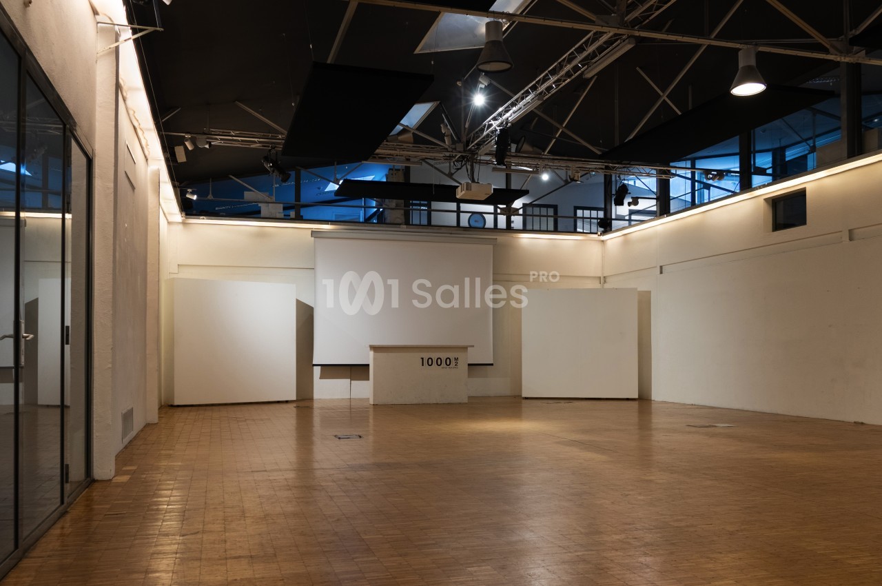 Salle vide avec parquet en bois, murs blancs, écran de projection et plafond industriel avec éclairage suspendu.