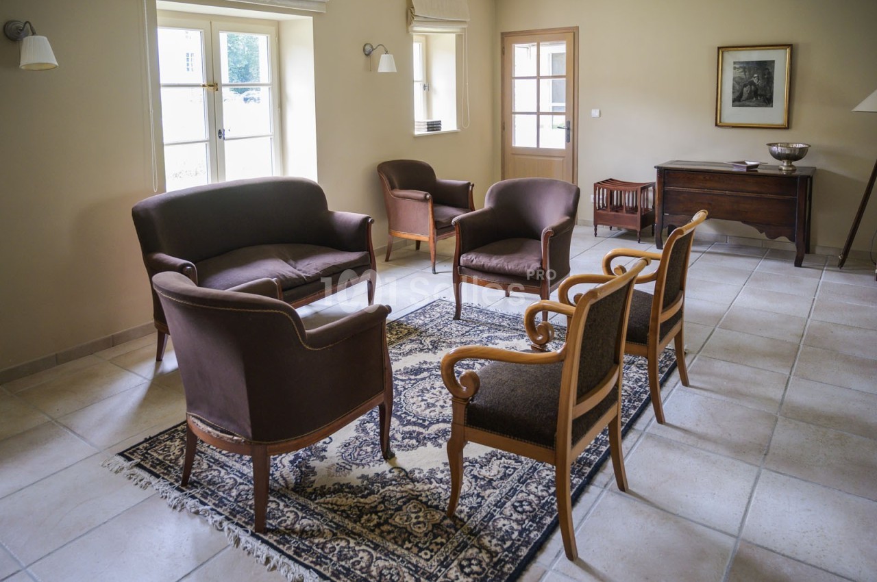 Salon avec fauteuils et canapé en bois et tissu, disposés autour d'un tapis, éclairé par des fenêtres à double battant.