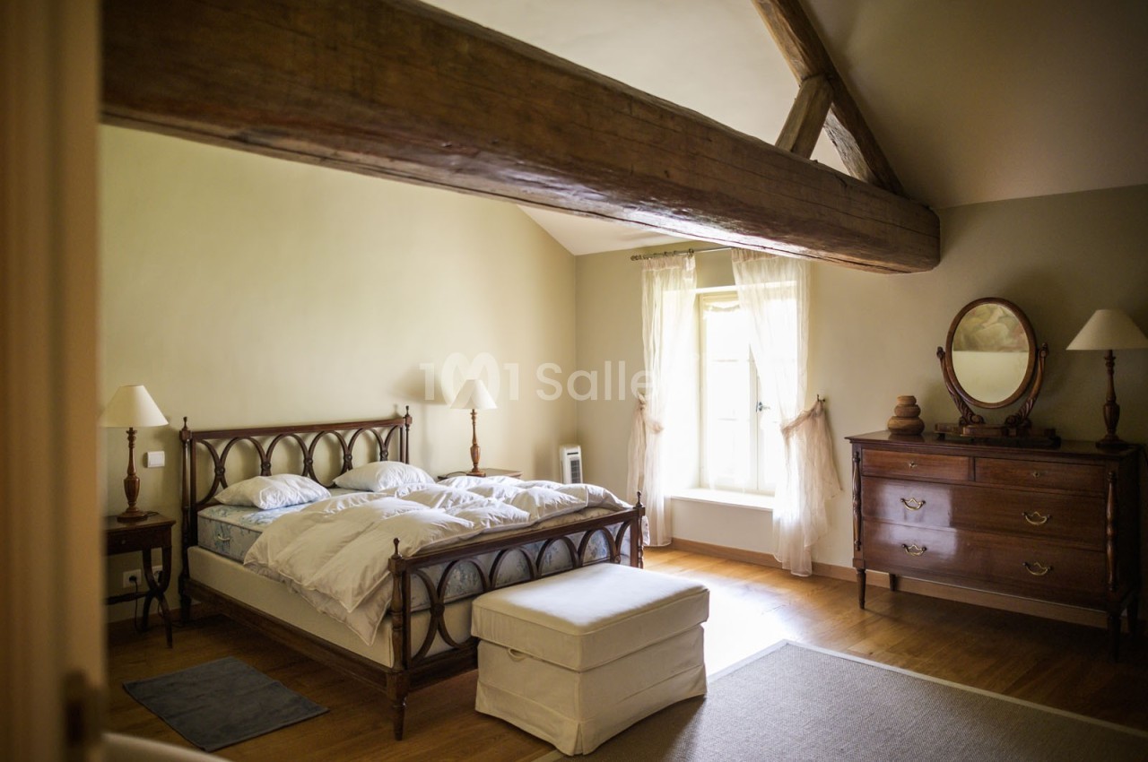 Chambre lumineuse avec lit double, poutre apparente, commode en bois, fauteuil et fenêtre avec rideaux blancs.