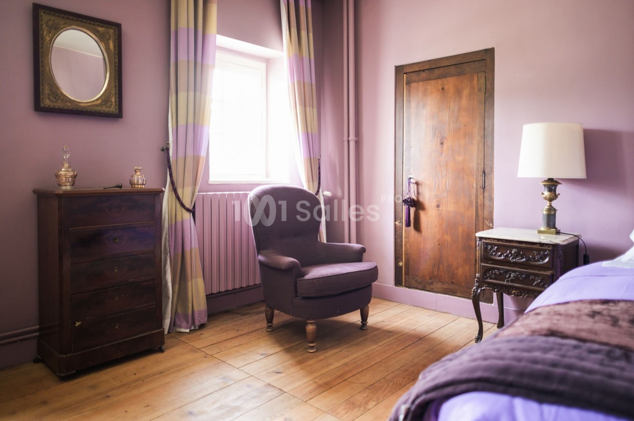 Chambre avec murs violets, fauteuil, commode en bois, lampe de chevet et fenêtre laissant entrer la lumière naturelle.