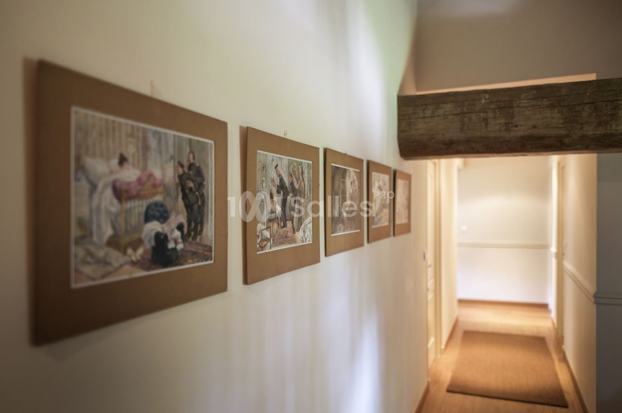 Tableaux encadrés accrochés sur un mur blanc dans un couloir éclairé par une lumière naturelle.