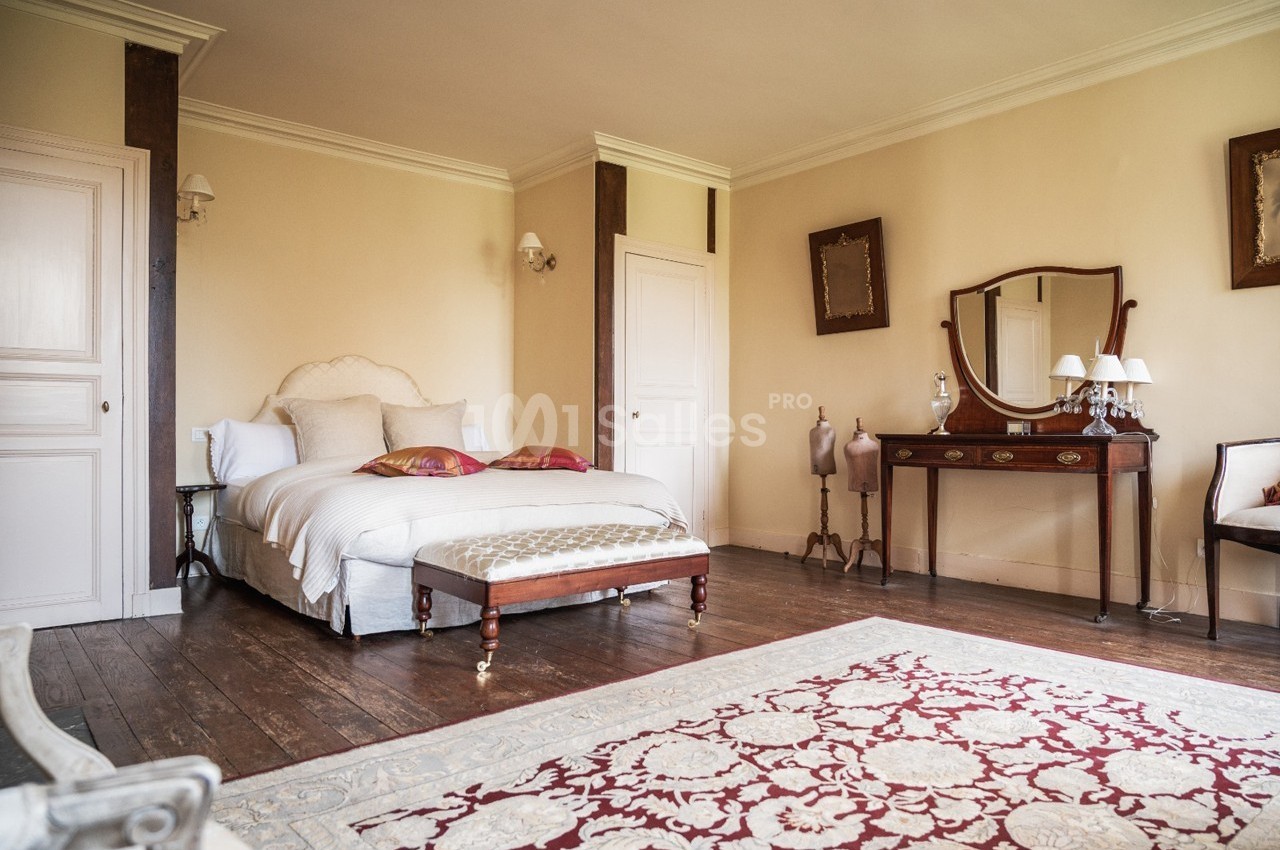 Chambre lumineuse avec lit double, mobilier ancien en bois, tapis rouge et beige, et murs de couleur crème.