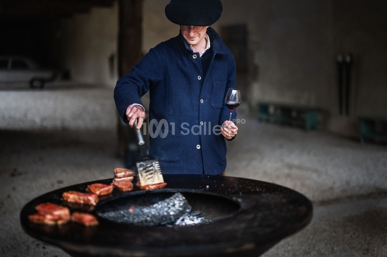 Homme en veste bleue cuisant des morceaux de viande sur un brasero, un verre de vin rouge à la main.