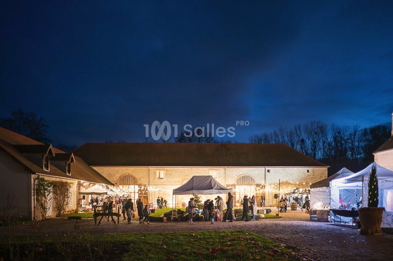 Cour d'une ferme illuminée en soirée, avec des stands, des lumières et des visiteurs rassemblés.