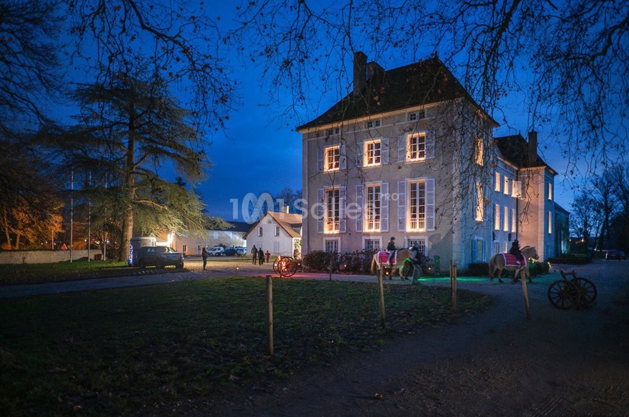 Façade éclairée d'un bâtiment ancien entouré d'arbres, avec des visiteurs et des calèches en soirée.