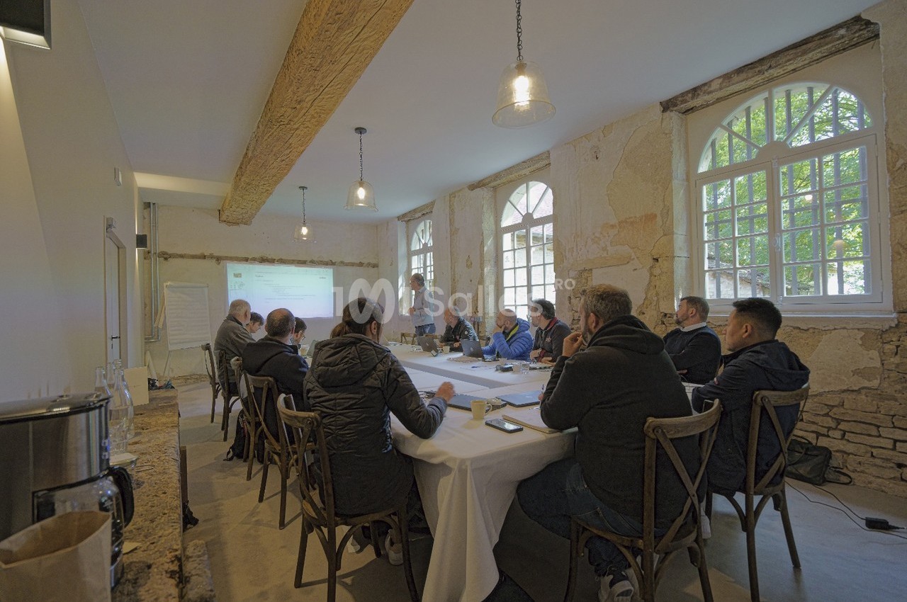 Un groupe de personnes assises autour d'une table dans une salle lumineuse, assistant à une présentation projetée au mur.