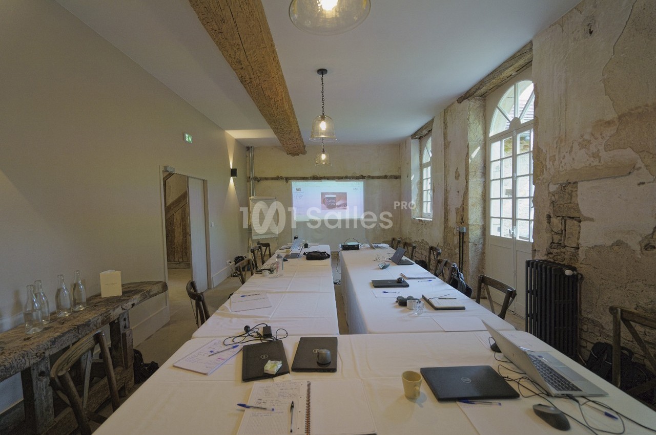 Salle de réunion rustique avec murs en pierre, grande table rectangulaire, ordinateurs portables et projecteur allumé.