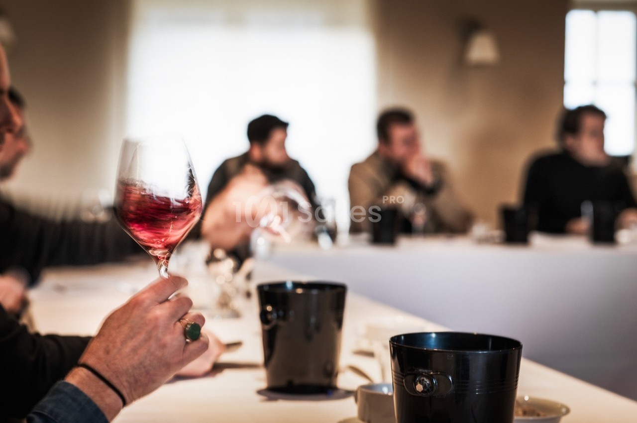 Une personne tient un verre de vin rouge lors d'une dégustation dans une salle avec plusieurs participants.