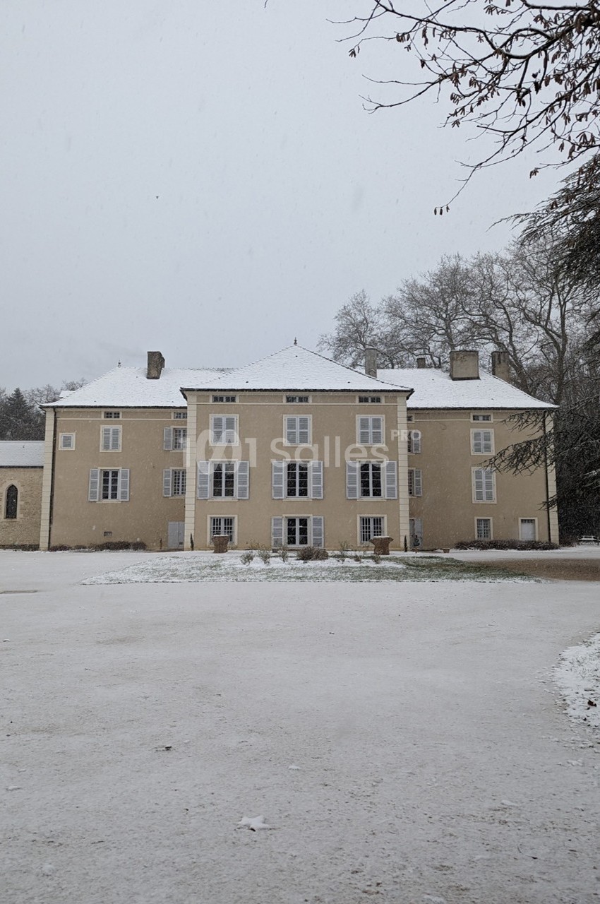 Façade d'un bâtiment ancien beige avec toit enneigé, entouré d'arbres en hiver.