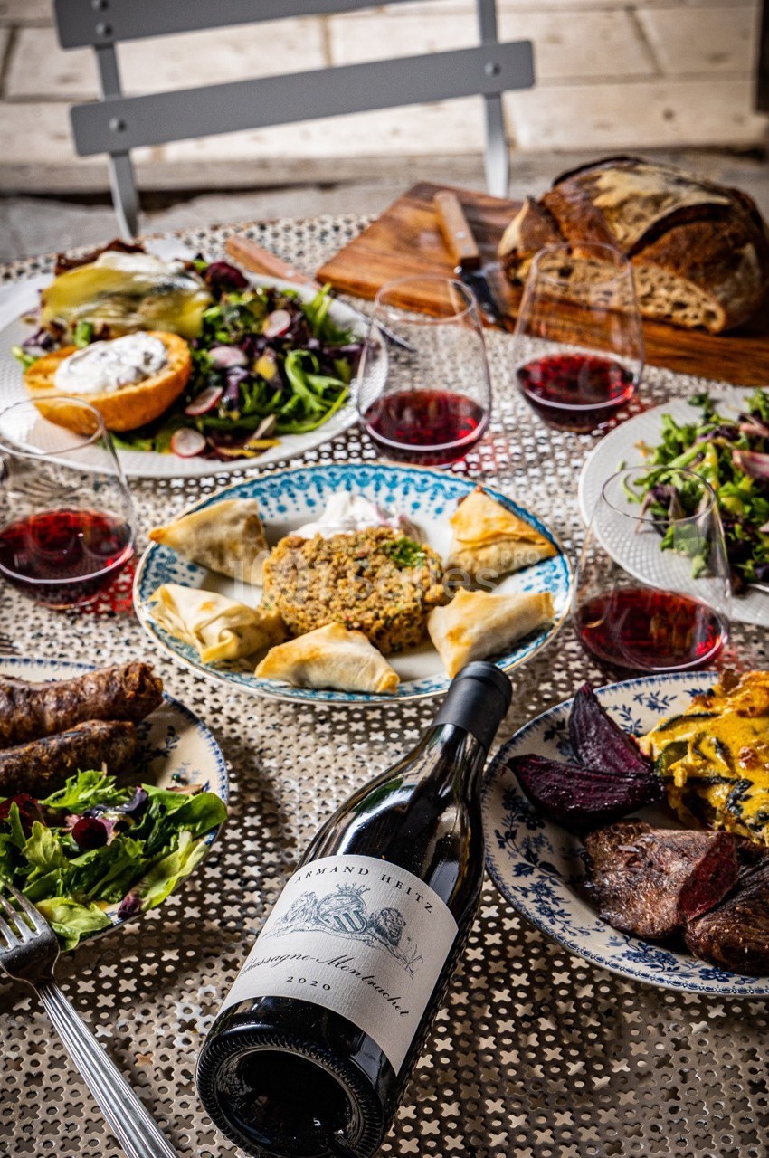 Table dressée avec des plats variés, du vin rouge, du pain, des salades et des mets cuisinés, sur une nappe ajourée.