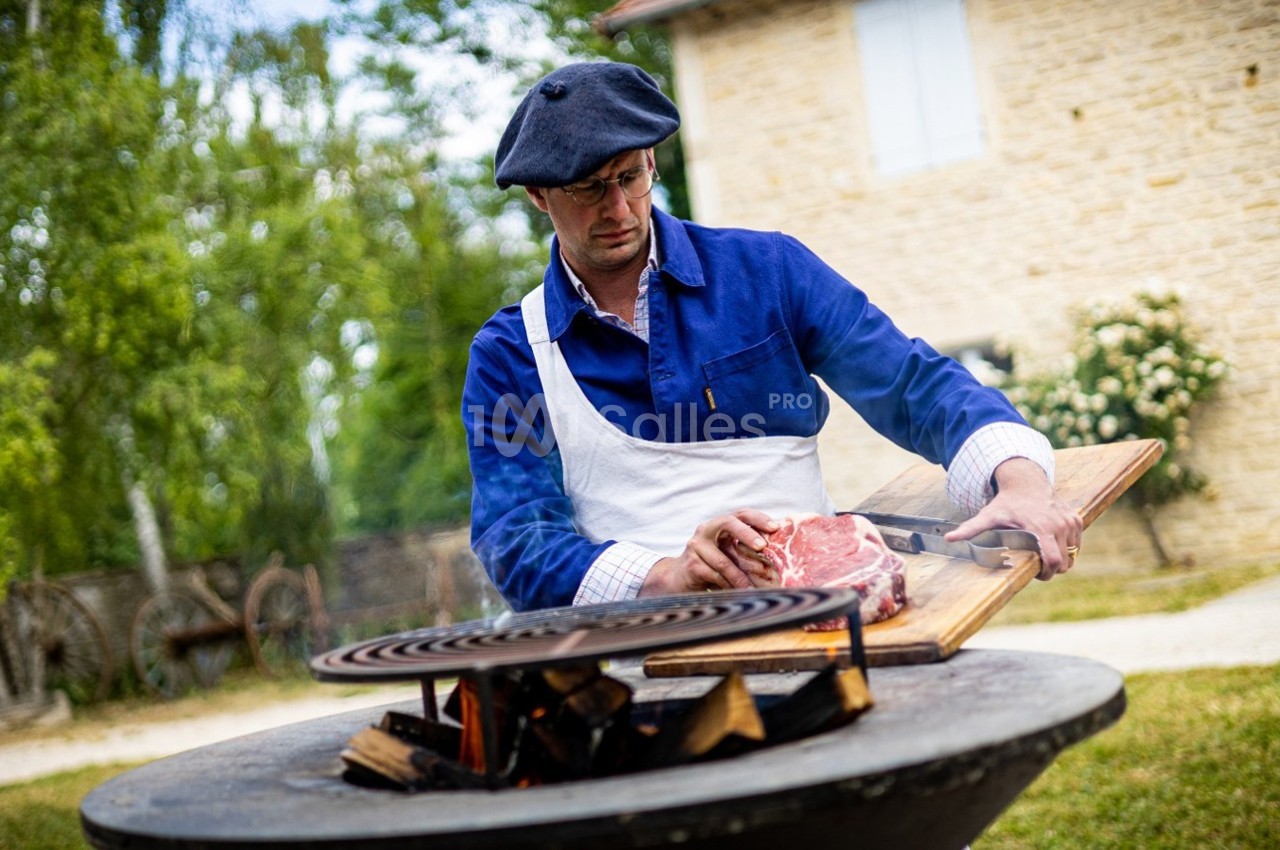 Homme en tablier préparant un morceau de viande sur une planche près d'un barbecue en plein air, devant une maison en pierre.