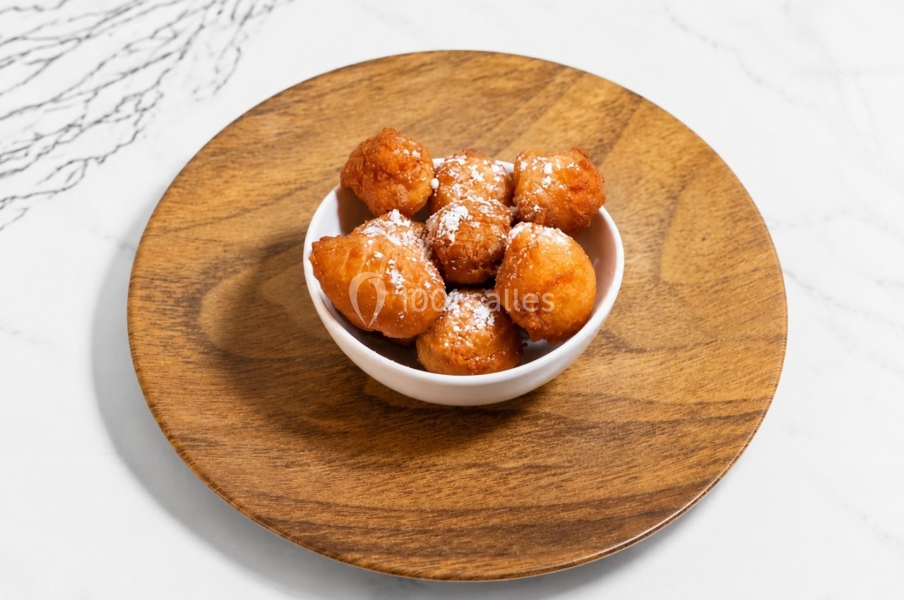 Beignets dorés saupoudrés de sucre glace, présentés dans un bol blanc sur une assiette en bois.