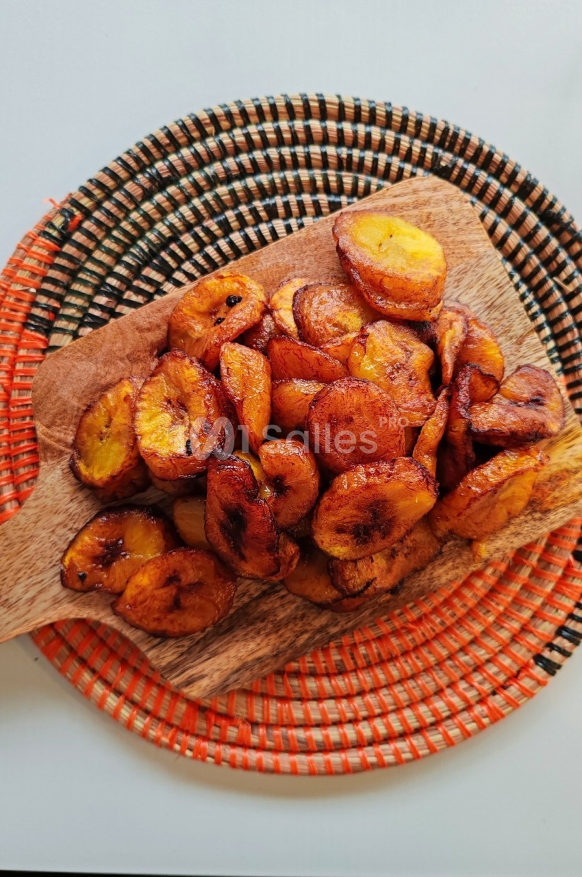 Tranches de bananes plantains frites disposées sur une planche en bois, posée sur un dessous de plat coloré.
