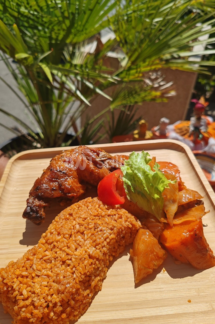 Assiette en bois contenant du riz rouge, du poulet grillé, des légumes et une garniture de salade, en extérieur ensoleillé.