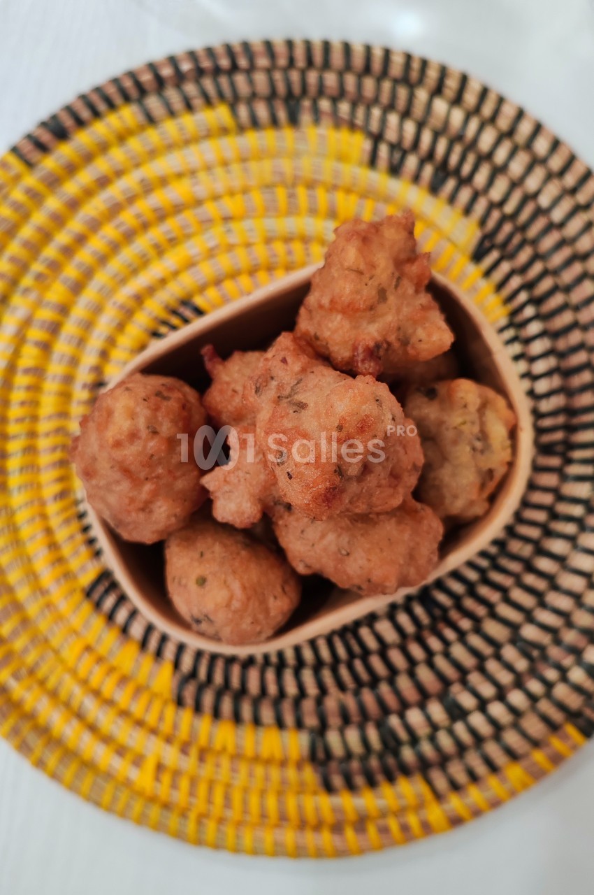 Beignets dorés servis dans un récipient en bois, posés sur un plateau tressé aux motifs jaunes et noirs.