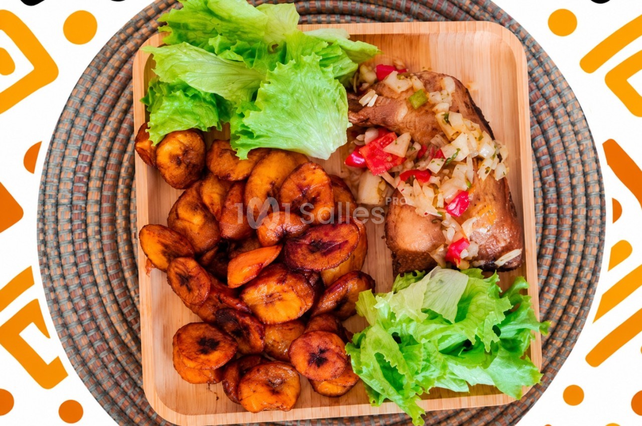 Assiette en bois contenant des rondelles de bananes plantains frites, du poulet garni de légumes et des feuilles de salade.