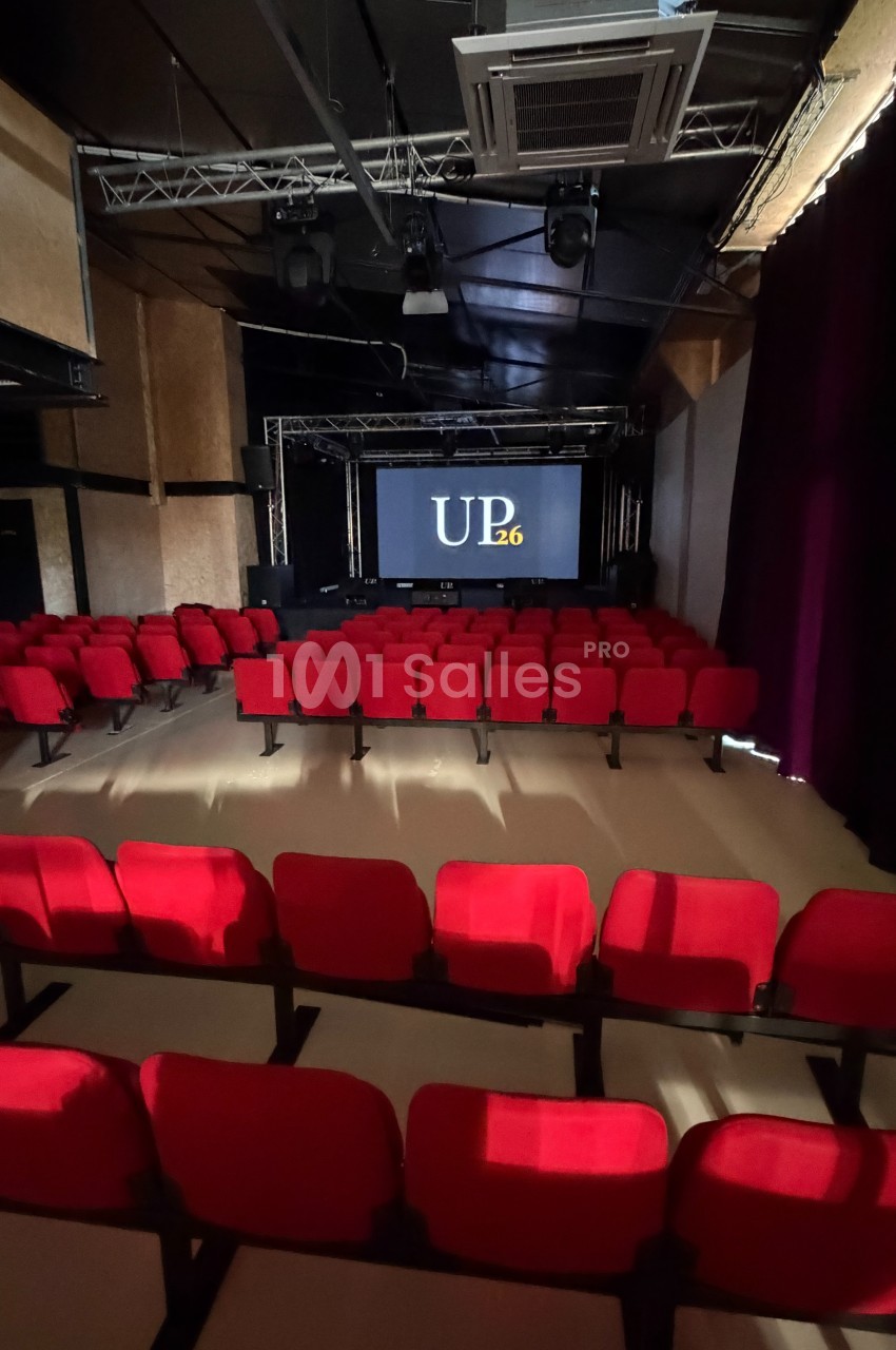 Salle de projection avec rangées de sièges rouges et un écran affichant ’UP26’ dans un espace sombre.