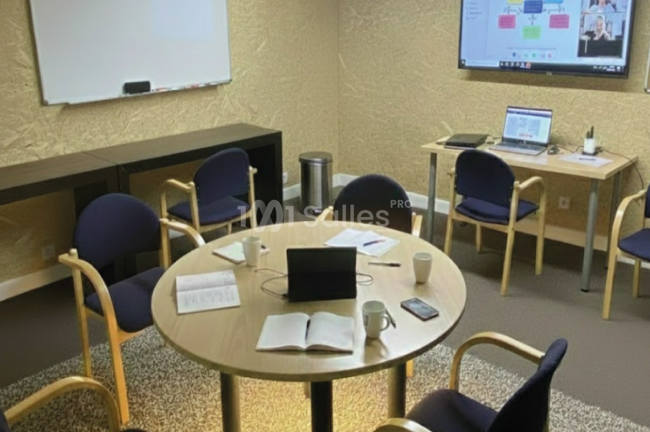 Salle de réunion avec table ronde, chaises, documents ouverts, écran de présentation et matériel informatique.