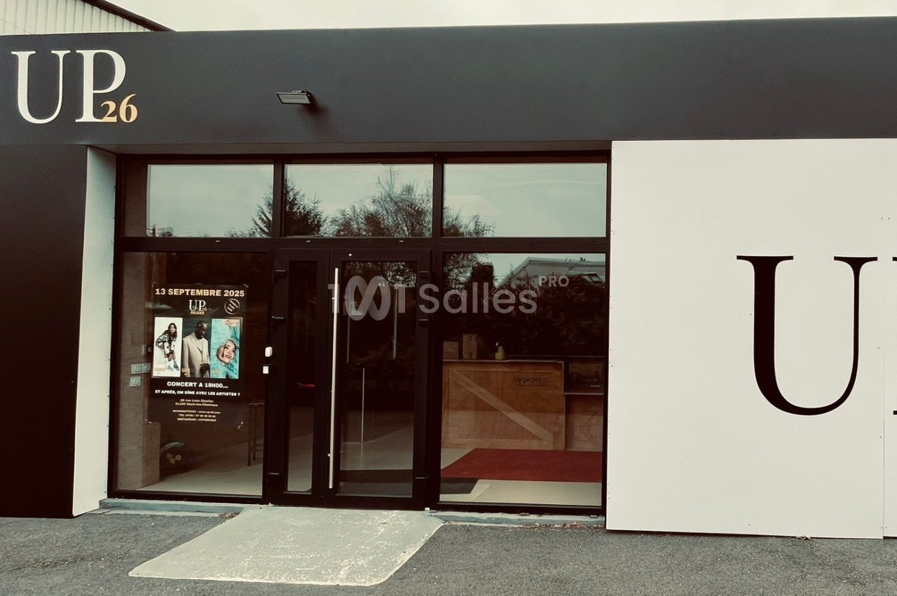 Façade d'un bâtiment avec une entrée vitrée, affiches promotionnelles sur la porte et enseigne ’UP26’ visible.