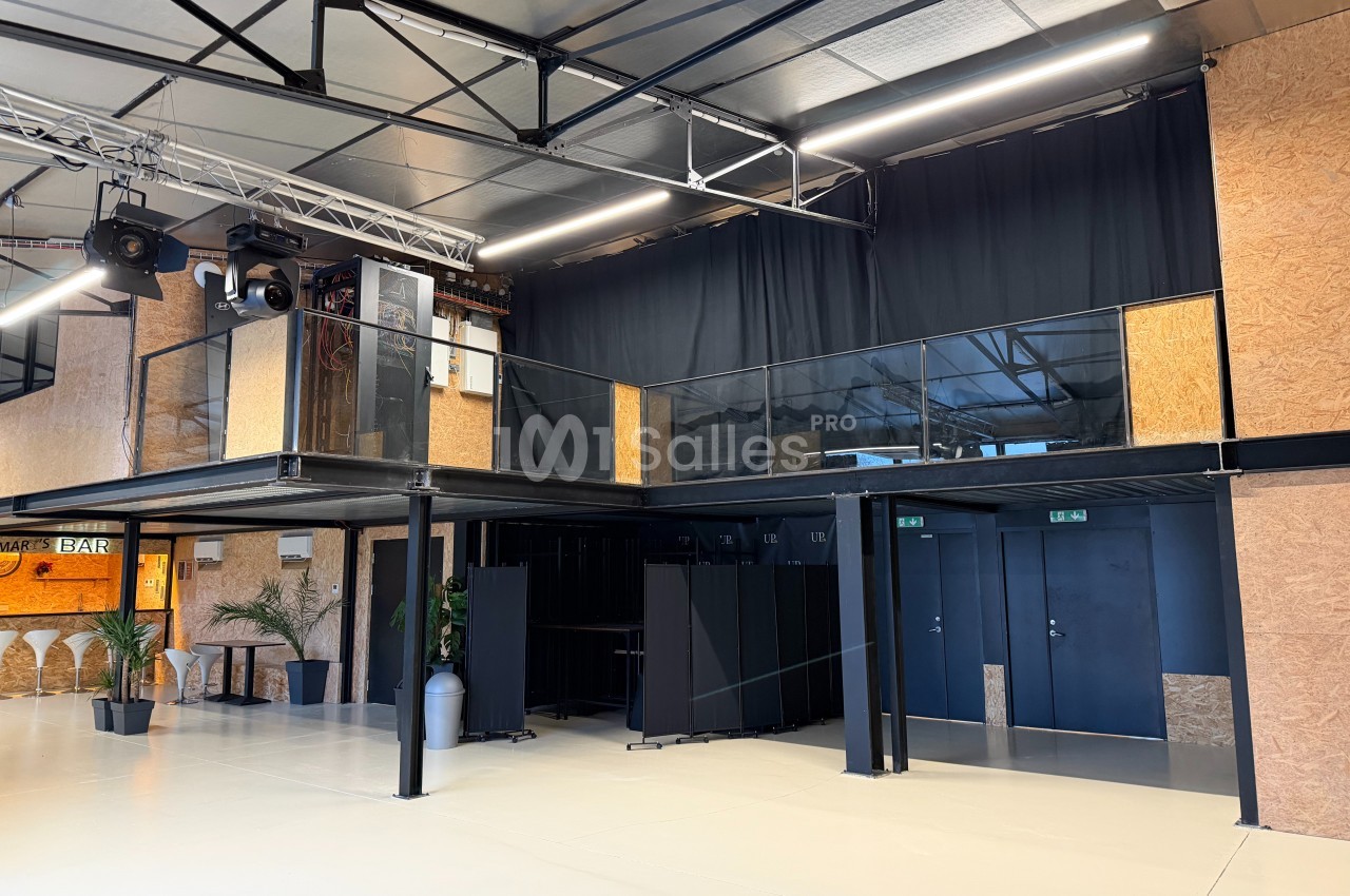 Salle moderne avec mezzanine en bois, rideaux noirs, éclairage suspendu et portes métalliques noires au rez-de-chaussée.