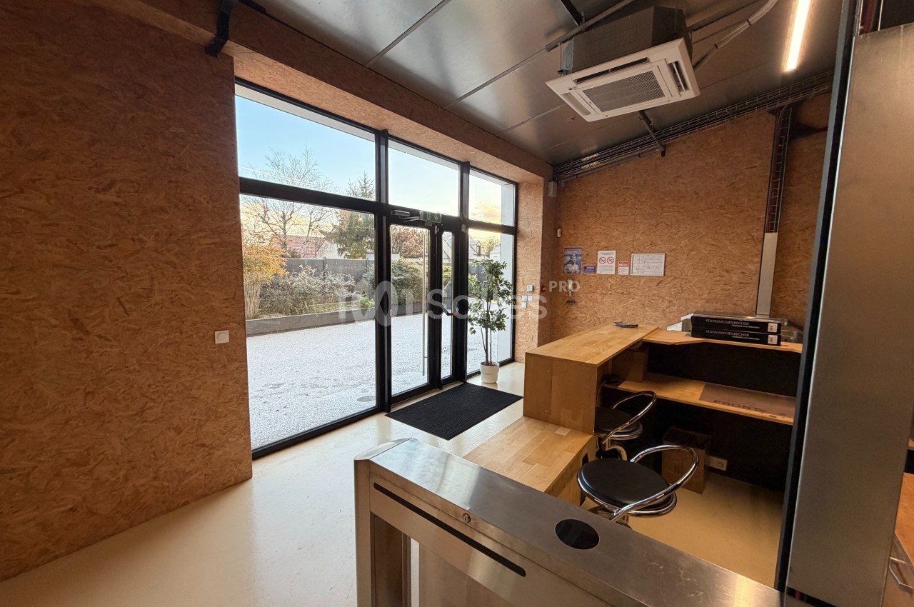 Entrée d'un bureau avec comptoir en bois, chaise, baie vitrée donnant sur un extérieur arboré et sol clair.