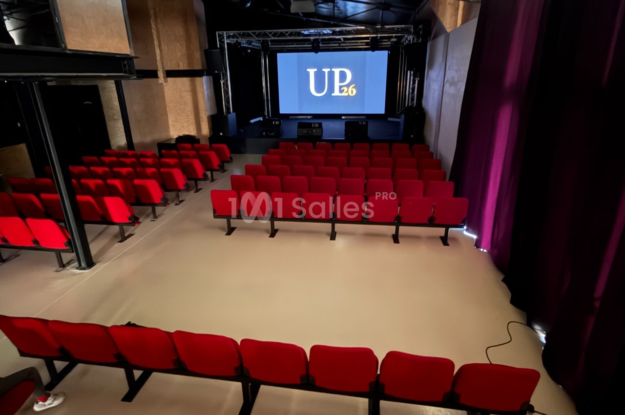 Salle de spectacle avec des rangées de sièges rouges face à une scène équipée d'un écran affichant ’UP26’.