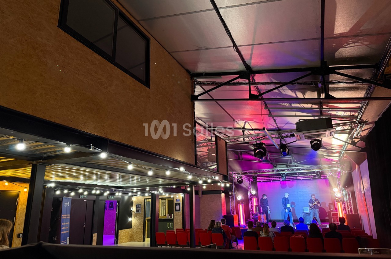 Salle de spectacle avec scène éclairée, artistes en représentation et public assis dans des fauteuils rouges.